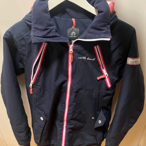 Mörkblå lätt fodrad skidjacka från North Bend - Säljer en snygg mörkblå lätt fodrad skidjacka från North Bend i mycket bra skick. Den har dragkedja i rosa, orangea och vita detaljer samt en logga på ärmen. 
