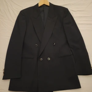 Svart kavaj från Oscar Jacobson - Elegant svart kavaj från Oscar Jacobson, tillverkad i Italien med Super 100's tyg från Loro Piana. Dubbelknäppt design med klassiska knappar och en stilren passform. Perfekt för formella tillfällen.                                 Storlek 48 - motsvara M