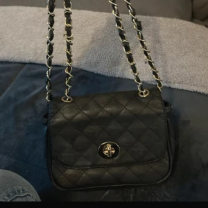 Svart quiltad axelväska - Snygg svart quiltad axelväska med kedjedetaljer på axelremmen och ett vridlås framtill. Perfekt för att ge en elegant touch till din outfit.