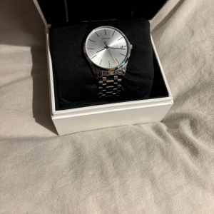 Silverfärgad klocka från Calvin Klein - Elegant silverfärgad klocka från Calvin Klein med stilren urtavla och datumvisning. Armbandet är i rostfritt stål och ger en klassisk look. Perfekt för dig som gillar tidlös design. Länkar med följer och kvitto. PRIS KAN SÄNKAS