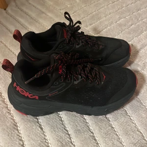 Svarta och röda sneakers från Hoka - Snygga svarta sneakers från Hoka med röda detaljer och snörning. Skorna har en robust sula med bra grepp, perfekt för träning eller vardagsbruk. Hoka-loggan syns tydligt på sidan. De är använda två ggr då det blev fel köp, fel storlek. 😊