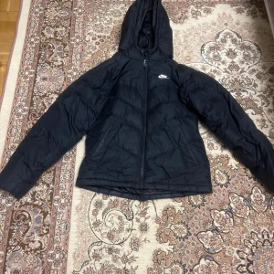 Svart dunjacka från Nike - Säljer en svart dunjacka från Nike med huva och dragkedja. Jackan har en klassisk pufferdesign och är perfekt för kyligare väder. Den har långa ärmar och en liten Nike-logga på bröstet. Size L för barn