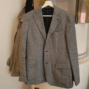 Grå kavaj från Gutteridge - Snygg grå kavaj från Gutteridge i 100% linne. Kavajen har ett diskret mönster och klassisk design med två knappar framtill. Storlek 48 herr.