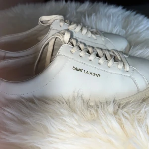 Vita sneakers från Saint Laurent - Säljer ett par  vita sneakers från Saint Laurent i skinn. De har klassisk snörning och märkeslogga på sidan. Dem är för små för mig därför jag väljer att sälja dem. Skorna har ett värde på ca 7000kr