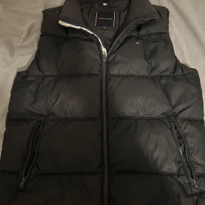 Svart dunväst från Tommy Hilfiger - Säljer en snygg svart dunväst från Tommy Hilfiger i mycket bra skick. Västen har en dragkedja framtill och två fickor med dragkedjor.