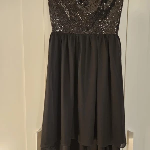 Svart paljettklänning - Snygg svart klänning med paljetter upptill och en flytande kjol. Perfekt för festliga tillfällen! Klänningen är axelbandslös och har en dragkedja i sidan för enkel påklädning. Den är i nytt skick och redo att glänsa på nästa fest.