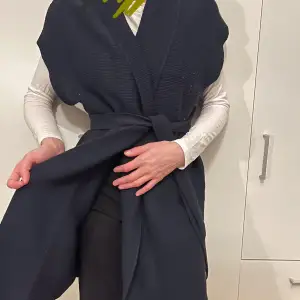 Säljer en snygg mörkblå stickad väst i mycket bra skick. Den har ett knytband i midjan och en stilren design som passar perfekt för höst och vår. Västen är ärmlös och har en normal passform, vilket gör den lätt att matcha med olika outfits.