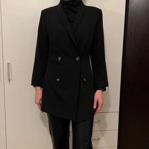 Svart kavaj med dubbelknäppning - Snygg svart kavaj i normal passform med dubbelknäppning framtill. Perfekt för både formella och semi-formella tillfällen. Kavajen har en klassisk design med två rader av knappar och är tillverkad i ett stilrent material.