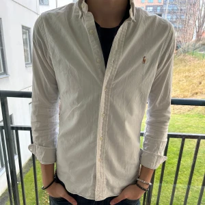 Vit skjorta från Ralph Lauren - Säljer en stilren vit skjorta från Ralph Lauren i slim fit. Skjortan är i stretchigt oxfordmaterial och har långa ärmar. Perfekt för både vardag och fest! Den har knappar framtill och en liten broderad logga på bröstet. Skjortan är i bra skick och redo för en ny ägare.