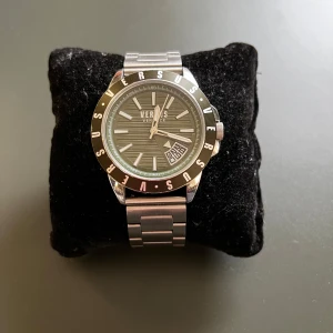 Versus Versace klocka i silver - Säljer en snygg Versus Versace klocka i silver och brittisk sport grön med ett stilrent armband och en cool urtavla. Klockan har tydliga index och datumvisning. Perfekt för både vardag och fest! Kommer med originalförpackning och extra armband. Ingår även ett autentiserings kort 