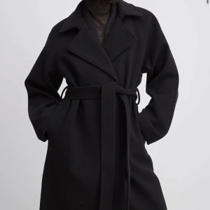 Svart kappa från NA-KD - Lång svart kappa från NA-KD med knytbälte i midjan. Helt ny med prislapp. Slutsåld på hemsidan