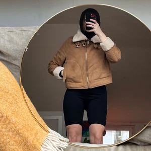 Brun mockajacka med pälsfoder från H&M - Säljer en snygg brun croppad mockajacka från H&M med mjukt pälsfoder. Jackan har en dragkedja och spännen vid kragen för extra stil. Perfekt för kyliga dagar med sin varma och bekväma design.