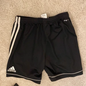 Svarta shorts från Adidas - Säljer ett par svarta Adidas shorts i mycket bra skick. De har de klassiska tre ränderna på sidan och är perfekta för träning eller fotboll. Materialet är lätt och andas bra, vilket gör dem bekväma att ha på sig under intensiva aktiviteter.