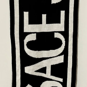 Svartvit Versace halsduk - Snygg och stilren halsduk i svart och vitt med ikonisk Versace text. Perfekt för att hålla sig varm och trendig under kyliga dagar. Passar både till vardags och speciella tillfällen.