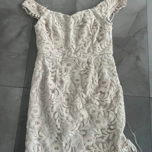 Beige spetsklänning - Elegant beige klänning med vackert blommigt spetsmönster. Klänningen har korta ärmar och en figurnära passform som framhäver silhuetten. Perfekt för speciella tillfällen.