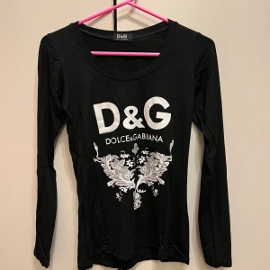 Svart långärmad topp från Dolce & Gabbana - Snygg svart långärmad topp från Dolce & Gabbana med vit logotyp och dekorativt mönster framtill. Perfekt för en stilren look.