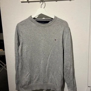 Grå tröja från Tommy Hilfiger - Säljer en stilren grå tröja från Tommy Hilfiger med rund hals och diskret logotyp på bröstet. Tröjan har långa ärmar och ribbade muddar vid ärmslut och nederkant. Perfekt för en avslappnad look. M