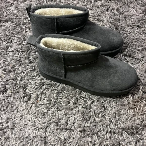 Grå boots med foder - Mysiga grå boots i mocka med varmt foder inuti. Perfekta för kalla dagar och håller fötterna varma och sköna. De har en enkel design utan snörning eller dragkedja, vilket gör dem lätta att ta på och av. Passar perfekt till både vardag och avslappnade tillfällen.