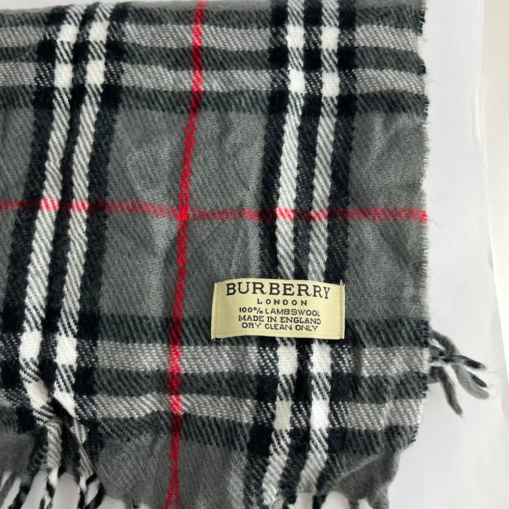 Säljer en klassisk grå rutig halsduk från Burberry i 100% lammull. Den har ett tidlöst mönster med inslag av svart, vitt och rött. Perfekt för att hålla sig varm under kyliga dagar. Fransen ger en extra touch av stil. Passar både höst och vinter. 🧣✨. Övrigt.