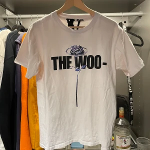 Vlone tee - Säljer en vit t-shirt från Vlone i bomull med tryck fram och bak. Framsidan har texten 'THE WOO-' med en ros, och baksidan har ett stort 'V' med blommönster. Perfekt för en avslappnad stil!