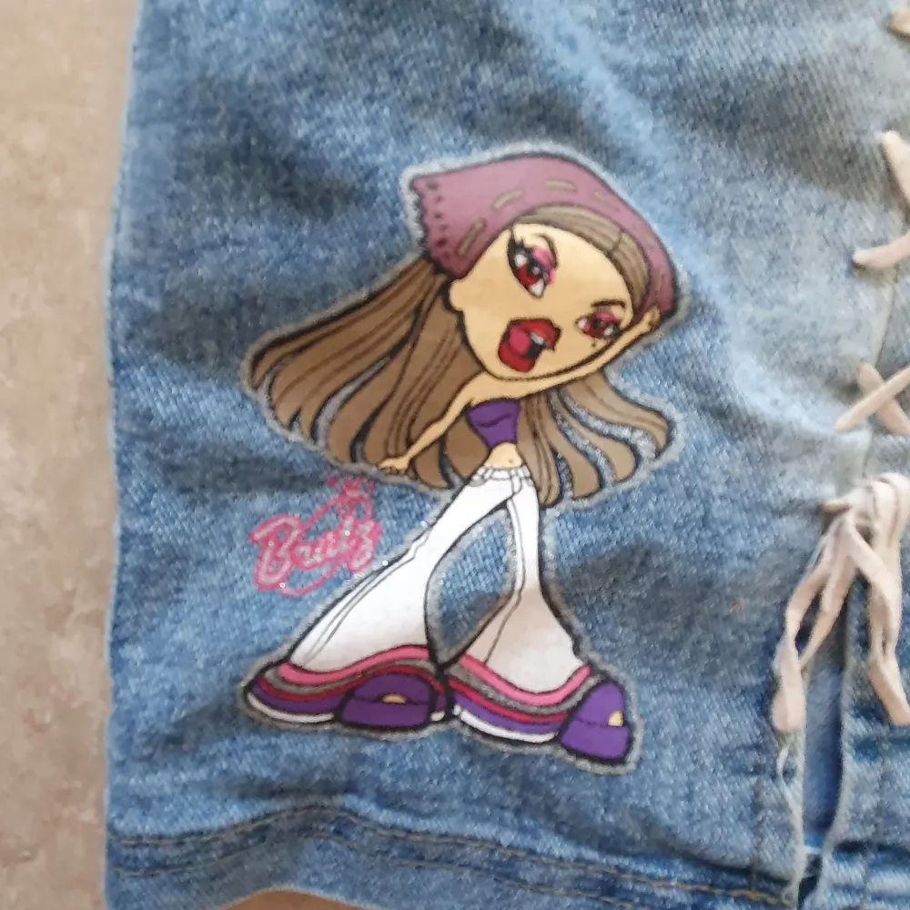 Säljer en supercool jeanskjol från Bratz med snörningsdetaljer på framsidan och en färgglad Bratz-figur på ena sidan. Kjolen har en klassisk blå jeansfärg och två bakfickor. Perfekt för att ge din outfit en retro touch!. Kjolar.