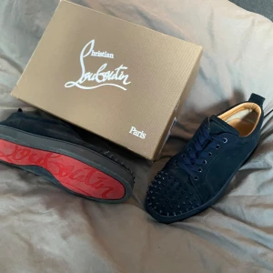 Christian Louboutin - Snygga svarta sneakers i mocka från Christian Louboutin med ikoniska röda sulor. Skorna har nitar på tån och snörning framtill. Perfekta för att ge en edgy touch till din stil.
