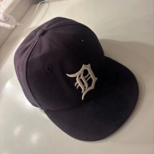 Svart keps från New Era - Säljer en svart keps från New Era, modell 59FIFTY. Den har en broderad vit bokstav på framsidan och är i storlek 6 3/4. Perfekt för både vardag och sportiga tillfällen. Kepsen är i bra skick och redo för nya äventyr!