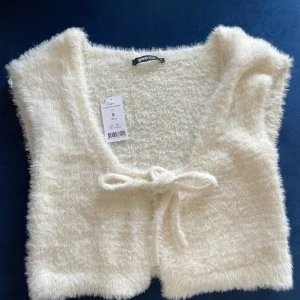 Vit fluffig väst från Gina Tricot - Superfin vit fluffig väst från Gina Tricot i storlek S. Den är gjord i ett mjukt och mysigt material och har en söt knytning framtill. Perfekt för att lägga till lite stil och värme till din outfit. Passar bra till både vardag och fest!