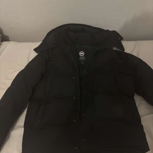 Svart dunjacka från Canada Goose - Säljer en svart dunjacka från Canada Goose. Jackan har en klassisk design med dragkedja och knappar framtill. Perfekt för kalla vinterdagar med sin varma och isolerande fyllning.