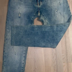 Blå jeans med slitningar - Säljer ett par blå jeans med coola slitningar och en klassisk femficksdesign. De har en knappgylf och en avslappnad passform som ger en skön stil. Perfekta för en avslappnad look.