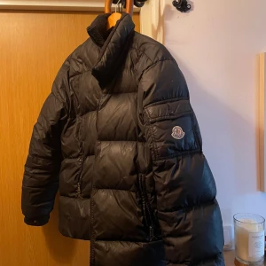 Svart dunjacka från Moncler - Säljer en snygg svart dunjacka från Moncler med hög krage och dragkedja. Jackan har en broderad logotyp på ärmen och praktiska fickor. Perfekt för kyliga dagar. Passar både tjej och kille storlek m men kanske L för tjejer. Dragkedjan på ena fickan är lite trög. Använd men fint skick. 