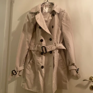 Beige trenchcoat från Zara - Snygg beige trenchcoat från Zara med dubbelknäppning och bälte i midjan. Jackan har långa ärmar och klassisk krage, perfekt för vår och höst. Stilsäker och tidlös design.