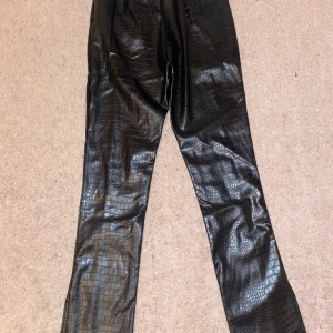 Svarta Gina Tricot fejkskinn byxor - Svarta fejkskinn byxor med ormskinnsmönster. Slits längs nedrebenet och dragskedja på ena höften. 