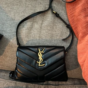 Svart axelväska från Saint Laurent - Snygg svart axelväska från Saint Laurent i skinn med quiltad design och guldiga detaljer. Väskan har en justerbar axelrem och ikonisk YSL-logga på framsidan. Perfekt för att ge en lyxig touch till din outfit. Den är köpt här på plick så har inget äkhetsbevis därav pris!