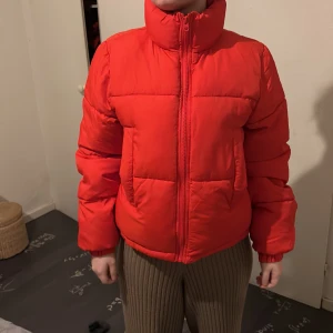 Röd pufferjacka från Gina Tricot - Säljer en snygg röd pufferjacka från Gina Tricot. Jackan har en hög krage och dragkedja framtill. Perfekt för kyliga dagar med sin varma och vadderade design. Ett måste för garderoben!