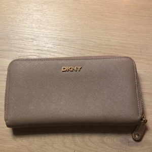 Beige plånbok från DKNY - Säljer en elegant beige plånbok från DKNY i mycket bra skick. Den har en praktisk dragkedja och flera fack för kort och kontanter. Perfekt för att hålla ordning på dina värdesaker med stil!