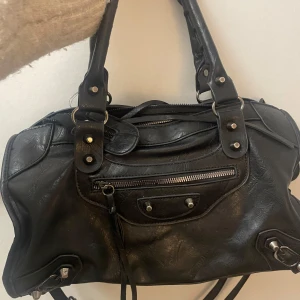 Svart axelväska i skinn - Intessekoll på min snygga svart axelväska i skinn med dragkedja och nitar. Perfekt för både vardag och fest. Väskan har en cool och stilren design med praktiska fack och detaljer.