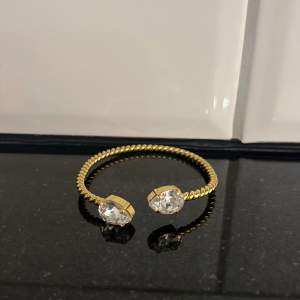 Snyggt guldarmband med en tvinnad design och två stora, gnistrande kristaller i ändarna. Perfekt för att ge lite extra bling till din outfit, oavsett om det är till vardags eller fest. Armbandet är justerbart och passar de flesta handleder. En riktig eye-catcher!