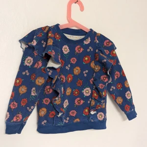 Blommig tröja med volanger från Minimarket - Superfin blå barn tröja från Minimarket med färgglatt blommönster. Den har långa ärmar och coola volanger