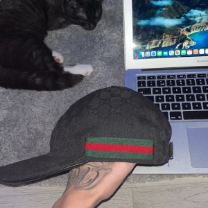 Svart keps från Gucci - Säljer en snygg svart keps från Gucci med det klassiska gröna och röda bandet. Kepsen är i mycket bra skick och har ett diskret mönster. Perfekt för att ge din outfit en lyxig touch. Kommer med originalförpackning.