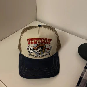 Oanvänd! Stetson keps - Stetson keps med texten 'Gambling Grifter' och ett spelkortsmotiv. Kepsen har en beige mesh-bakdel och en mörkblå. Justerbar passform för att sitta bekvämt på huvudet. Endast testad! Kontakta vid frågor. MVH M.sellout1337. Fläckfri de som syns på bilden som ser ut som fläckar är enbart damm!