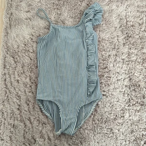 Randig baddräkt med volang - Säljer en superfin randig baddräkt i blått och vitt. Den har en unik design med en axel och en söt volangdetalj på sidan. Perfekt för sommarens alla badäventyr!. Den är 120 cm lång! 🥰