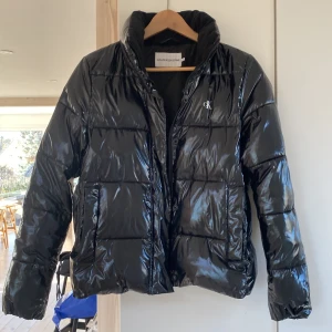 Svart pufferjacka från Calvin Klein - Säljer en snygg svart pufferjacka från Calvin Klein i mycket bra skick. Jackan är perfekt för kalla dagar med sin glansiga yta och varma vaddering. Den har en diskret CK-logga på bröstet och stängs med dragkedja. Passar perfekt för både höst och vinter! Finns även luva som går att ta av och på