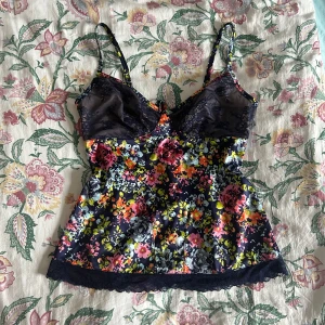 Blommig spetslinne - Säljer ett superfint blommigt linne med spetsdetaljer. Det har tunna axelband och en vacker blommig design i färger som rosa, blå och orange. Linnet har en snörning i ryggen.