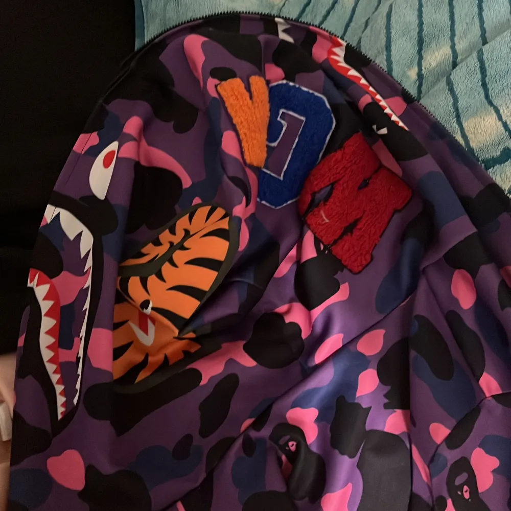 Säljer en cool hoodie från BAPE med ett unikt hajmotiv på luvan. Den har ett färgglatt camouflagemönster i lila, rosa och svart. Hoodien är långärmad med ribbade muddar och dragkedja. Perfekt för att sticka ut i mängden! Mycket bra skick priset kan diskuteras . Jackor.