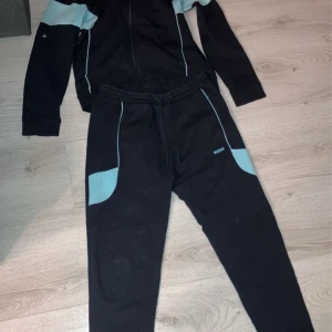 Svart och ljusblå träningsset från Boss - Säljer ett snyggt och bekvämt träningsset från Boss i svart med ljusblå detaljer. Setet består av en långärmad tröja med dragkedja och matchande byxor. Perfekt för träning eller en avslappnad dag. Passar bra till höst och vår.