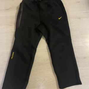 Svarta NOCTA byxor från Nike - Säljer ett par svarta NOCTA byxor från Nike som passar både M och L. Byxorna har en snygg gul logga och är perfekta för en avslappnad stil. De har snörning i midjan för bästa passform. Perfekta för både vardag och träning! Passar 