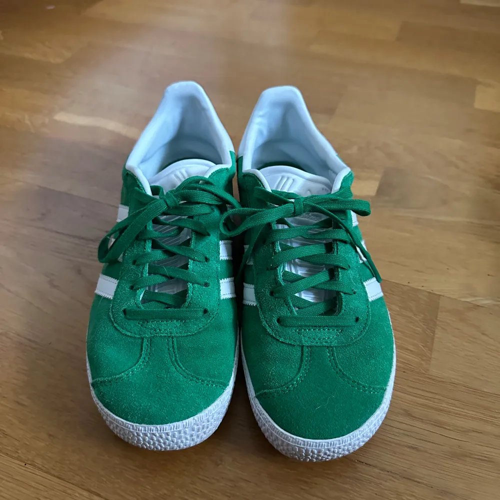 Säljer ett par snygga gröna Adidas Gazelle sneakers i mocka. De har vita detaljer och klassiska tre ränder på sidan. Skorna är i mycket bra skick och perfekta för både vardag och fest. Är perfekta för att lyfta en tråkig outfit. Använda endast fåtal gånger. Skor.