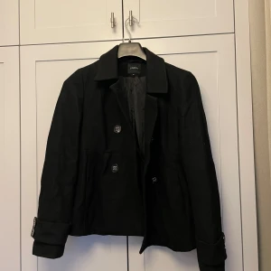 Kort kappa  - Snygg svart kappa från Lindex i mycket bra skick. Den har en klassisk design med dubbelknäppning och dekorativa knappar på ärmarna. Storlek dam M.