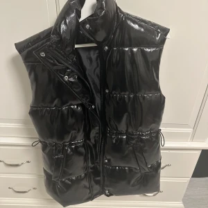 Svart glansig dunväst - Snygg svart glansig dunväst i mycket bra skick. Perfekt för höst och vinter med dragkedja och knappar framtill. Västen har en quiltad design och är normal i passformen. Den är både stilren och praktisk för kyligare dagar. Storlek XXS, men mer åt XS. Kan dessutom töjas ut lite till S vid kanterna 